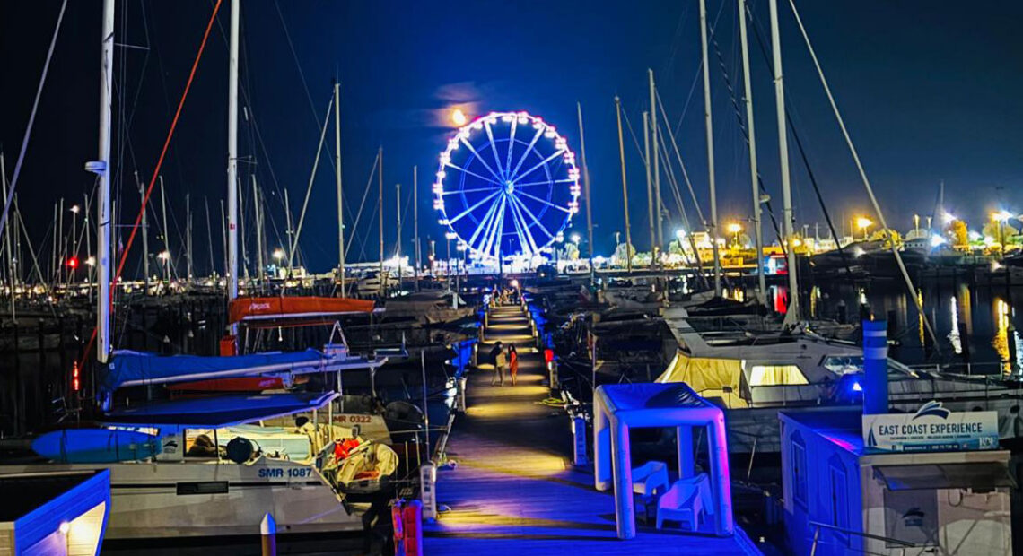 Veduta notturna del porto di Rimini con la ruota panoramica illuminata riflessa sul mare.