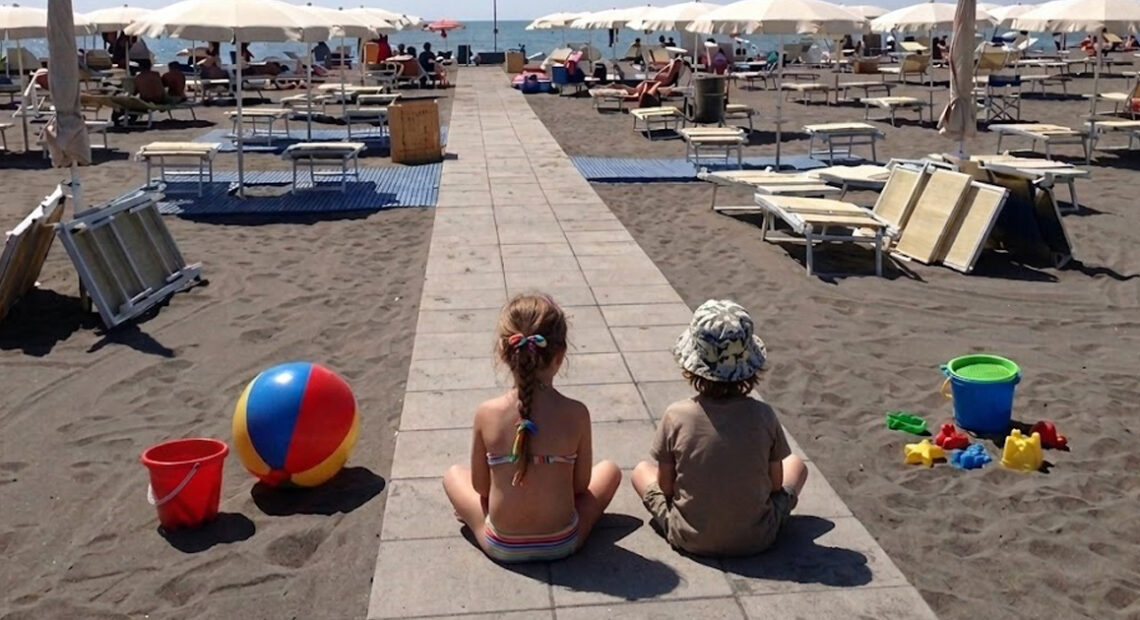 Due bambini seduti sulla passerella di uno stabilimento balneare a Montalto di Castro con giochi da spiaggia e ombrelloni.