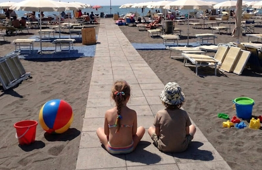 Due bambini seduti sulla passerella di uno stabilimento balneare a Montalto di Castro con giochi da spiaggia e ombrelloni.