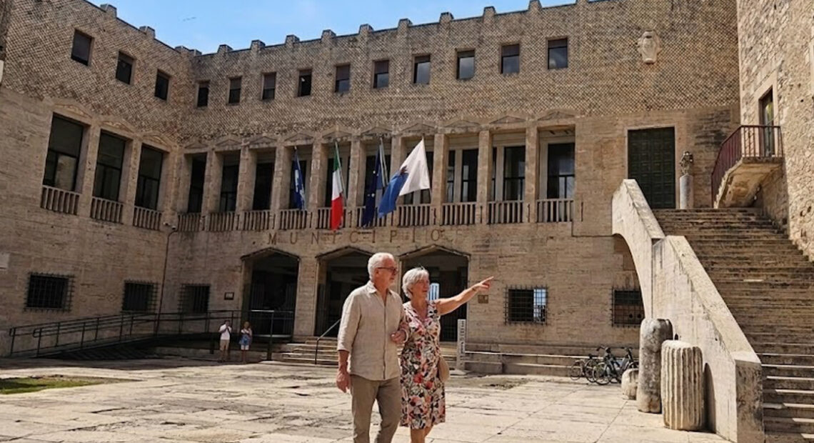 Facciata esterna del Palazzo Comunale di Terracina con bandiere istituzionali esposte sopra l'ingresso e persone nella piazza antistante.