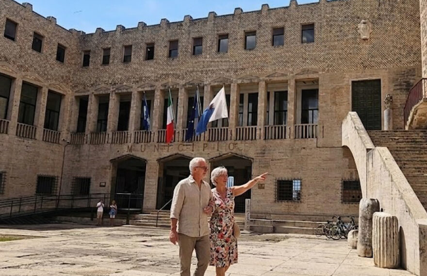Facciata esterna del Palazzo Comunale di Terracina con bandiere istituzionali esposte sopra l'ingresso e persone nella piazza antistante.