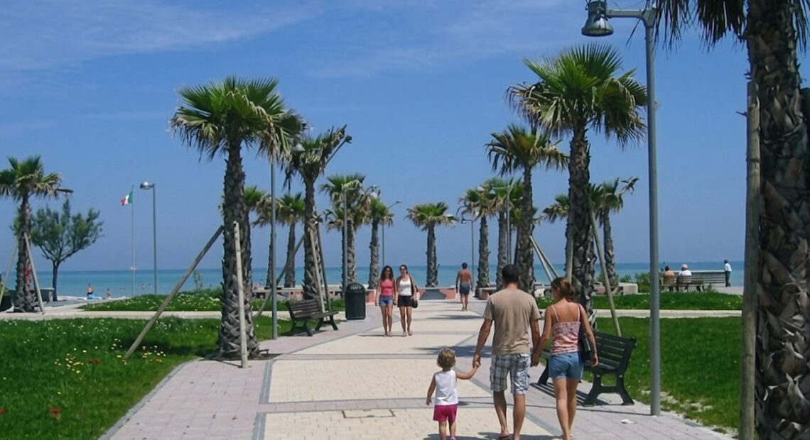 Il lungomare pedonale e ciclabile di Lido di Fermo con palme, aree verdi e turisti che passeggiano verso la spiaggia.