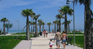 Il lungomare pedonale e ciclabile di Lido di Fermo con palme, aree verdi e turisti che passeggiano verso la spiaggia.