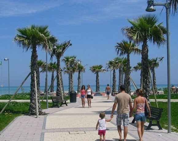 Il lungomare pedonale e ciclabile di Lido di Fermo con palme, aree verdi e turisti che passeggiano verso la spiaggia.
