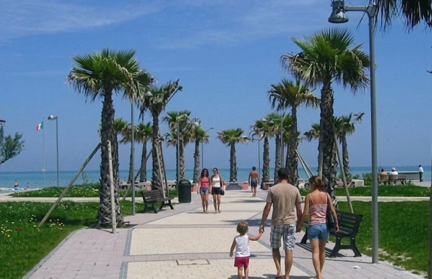 Il lungomare pedonale e ciclabile di Lido di Fermo con palme, aree verdi e turisti che passeggiano verso la spiaggia.