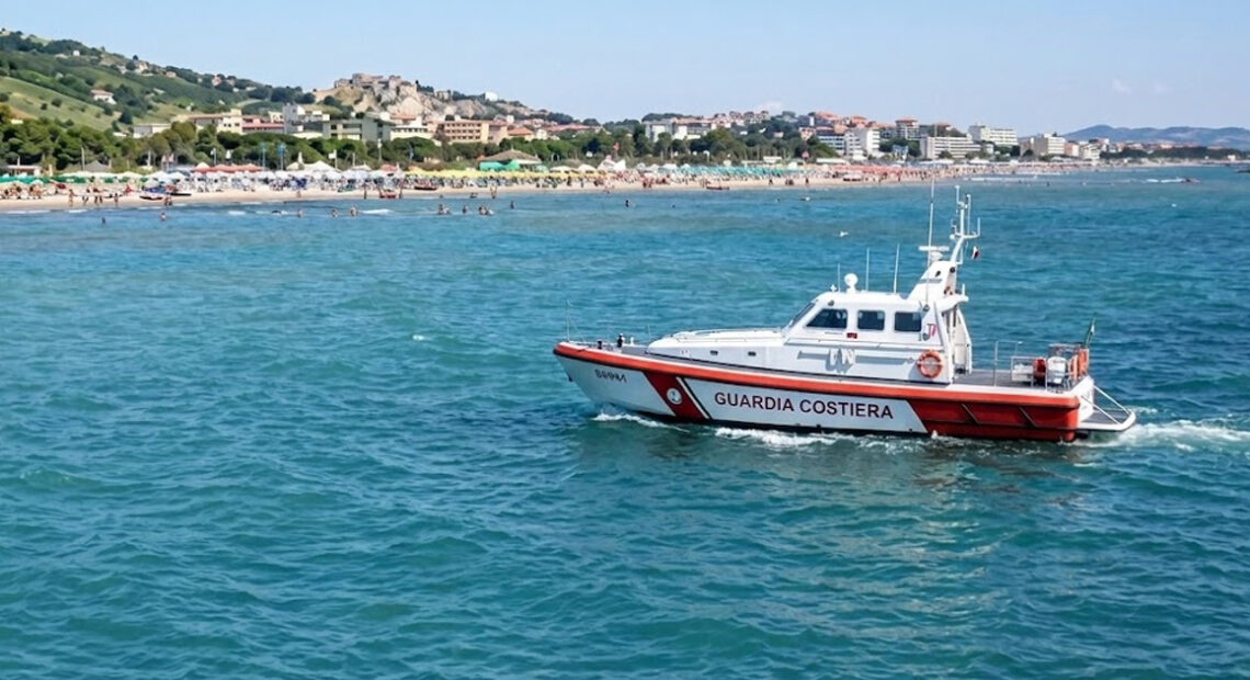 Motonave della Guardia Costiera in navigazione sottocosta davanti a una spiaggia affollata con stabilimenti balneari