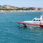 Motonave della Guardia Costiera in navigazione sottocosta davanti a una spiaggia affollata con stabilimenti balneari