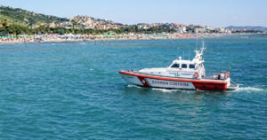 Motonave della Guardia Costiera in navigazione sottocosta davanti a una spiaggia affollata con stabilimenti balneari