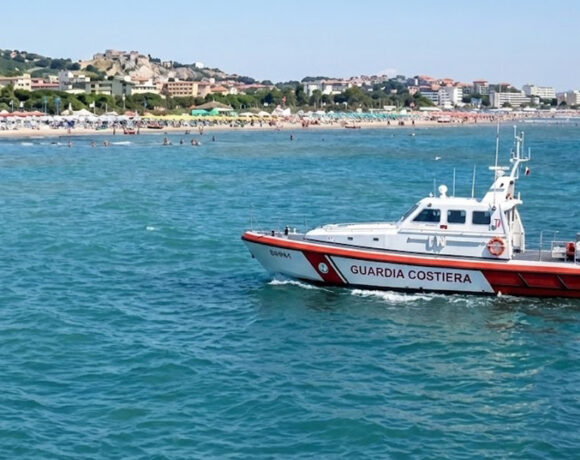 Motonave della Guardia Costiera in navigazione sottocosta davanti a una spiaggia affollata con stabilimenti balneari