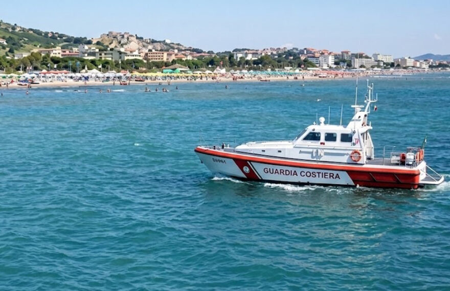 Motonave della Guardia Costiera in navigazione sottocosta davanti a una spiaggia affollata con stabilimenti balneari