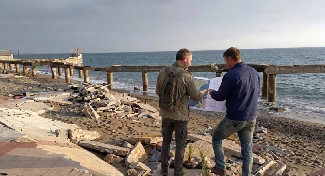 Due tecnici analizzano una mappa su una spiaggia colpita da forte erosione costiera con pavimentazione crollata.