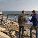 Due tecnici analizzano una mappa su una spiaggia colpita da forte erosione costiera con pavimentazione crollata.