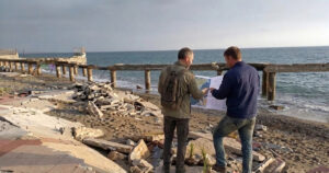 Due tecnici analizzano una mappa su una spiaggia colpita da forte erosione costiera con pavimentazione crollata.