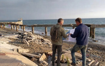 Due tecnici analizzano una mappa su una spiaggia colpita da forte erosione costiera con pavimentazione crollata.