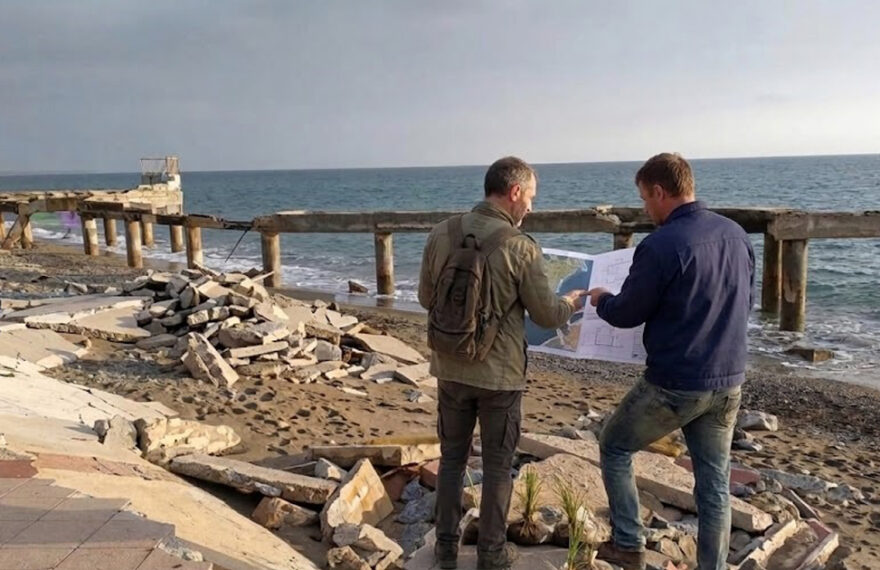 Due tecnici analizzano una mappa su una spiaggia colpita da forte erosione costiera con pavimentazione crollata.