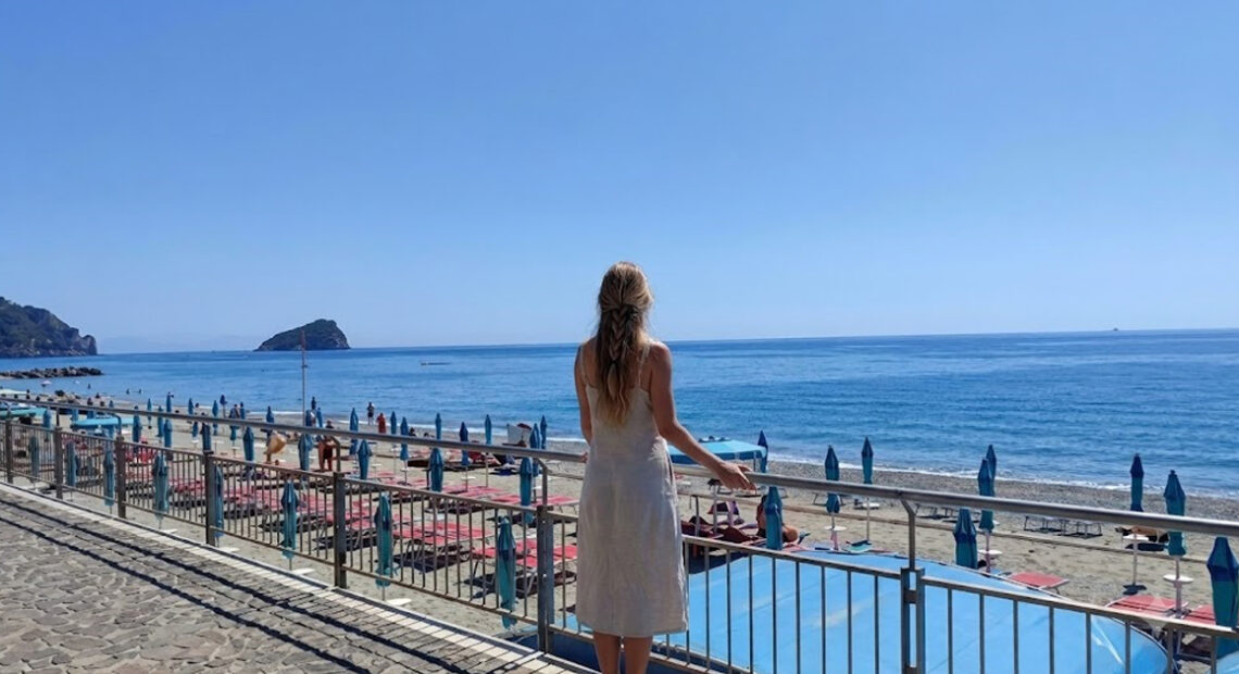 Una donna di spalle osserva il litorale e gli stabilimenti balneari di Spotorno con l'isola di Bergeggi sullo sfondo.