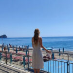 Una donna di spalle osserva il litorale e gli stabilimenti balneari di Spotorno con l'isola di Bergeggi sullo sfondo.