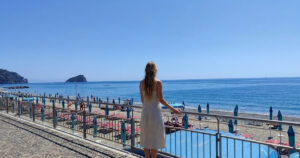 Una donna di spalle osserva il litorale e gli stabilimenti balneari di Spotorno con l'isola di Bergeggi sullo sfondo.