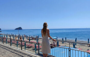 Una donna di spalle osserva il litorale e gli stabilimenti balneari di Spotorno con l'isola di Bergeggi sullo sfondo.