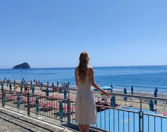 Una donna di spalle osserva il litorale e gli stabilimenti balneari di Spotorno con l'isola di Bergeggi sullo sfondo.