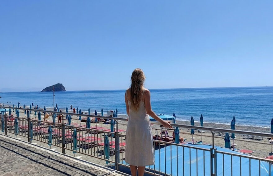 Una donna di spalle osserva il litorale e gli stabilimenti balneari di Spotorno con l'isola di Bergeggi sullo sfondo.