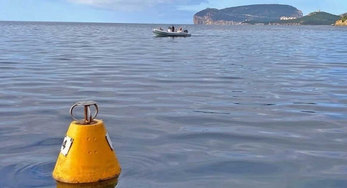 Una boa gialla conica per l'ormeggio ecologico che galleggia sulle acque calme dell'Area Marina Protetta di Capo Caccia, con un gommone in transito e il promontorio di Capo Caccia sullo sfondo.