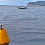 Una boa gialla conica per l'ormeggio ecologico che galleggia sulle acque calme dell'Area Marina Protetta di Capo Caccia, con un gommone in transito e il promontorio di Capo Caccia sullo sfondo.