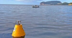 Una boa gialla conica per l'ormeggio ecologico che galleggia sulle acque calme dell'Area Marina Protetta di Capo Caccia, con un gommone in transito e il promontorio di Capo Caccia sullo sfondo.