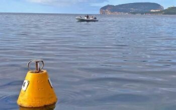 Una boa gialla conica per l'ormeggio ecologico che galleggia sulle acque calme dell'Area Marina Protetta di Capo Caccia, con un gommone in transito e il promontorio di Capo Caccia sullo sfondo.