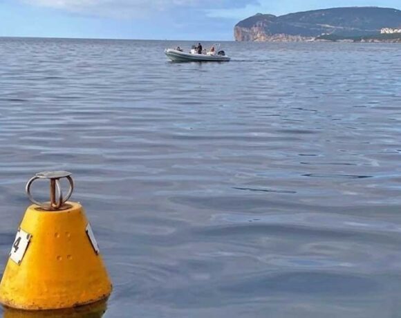 Una boa gialla conica per l'ormeggio ecologico che galleggia sulle acque calme dell'Area Marina Protetta di Capo Caccia, con un gommone in transito e il promontorio di Capo Caccia sullo sfondo.