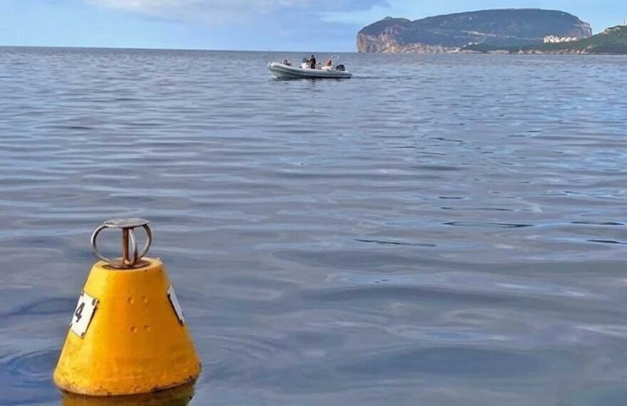 Una boa gialla conica per l'ormeggio ecologico che galleggia sulle acque calme dell'Area Marina Protetta di Capo Caccia, con un gommone in transito e il promontorio di Capo Caccia sullo sfondo.