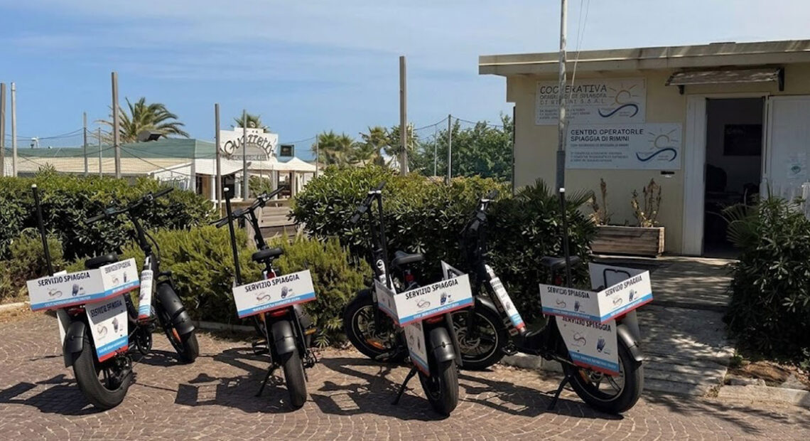 Una fila di biciclette elettriche e-bike nere con cartelli "Servizio Spiaggia" parcheggiate davanti alla sede della Cooperativa Operatori di Spiaggia di Rimini.