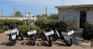Una fila di biciclette elettriche e-bike nere con cartelli "Servizio Spiaggia" parcheggiate davanti alla sede della Cooperativa Operatori di Spiaggia di Rimini.