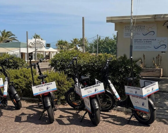 Una fila di biciclette elettriche e-bike nere con cartelli "Servizio Spiaggia" parcheggiate davanti alla sede della Cooperativa Operatori di Spiaggia di Rimini.