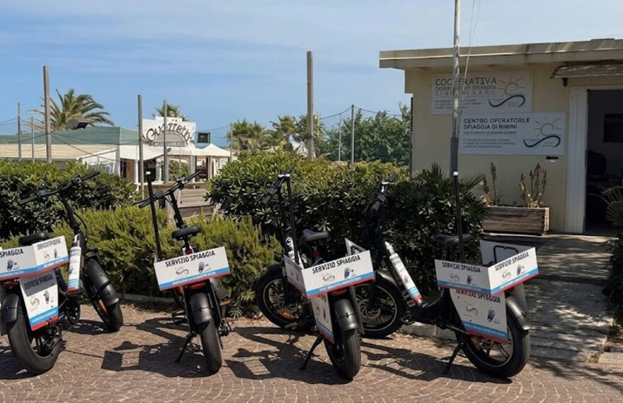 Una fila di biciclette elettriche e-bike nere con cartelli "Servizio Spiaggia" parcheggiate davanti alla sede della Cooperativa Operatori di Spiaggia di Rimini.