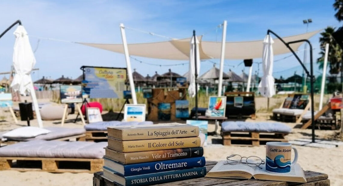 Libri e occhiali su un tavolo di legno in spiaggia a Pescara con quadri e lettini sullo sfondo per la rassegna Cultura al sole.