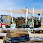 Libri e occhiali su un tavolo di legno in spiaggia a Pescara con quadri e lettini sullo sfondo per la rassegna Cultura al sole.