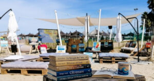 Libri e occhiali su un tavolo di legno in spiaggia a Pescara con quadri e lettini sullo sfondo per la rassegna Cultura al sole.