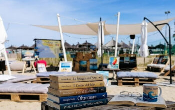 Libri e occhiali su un tavolo di legno in spiaggia a Pescara con quadri e lettini sullo sfondo per la rassegna Cultura al sole.
