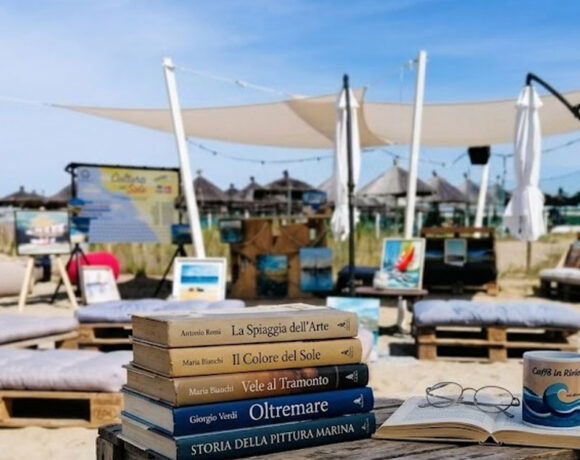 Libri e occhiali su un tavolo di legno in spiaggia a Pescara con quadri e lettini sullo sfondo per la rassegna Cultura al sole.