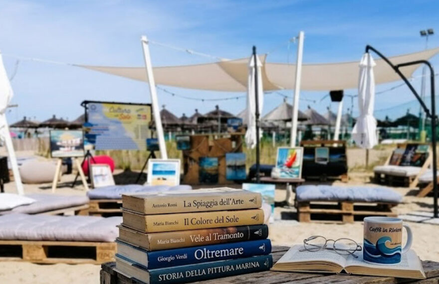 Libri e occhiali su un tavolo di legno in spiaggia a Pescara con quadri e lettini sullo sfondo per la rassegna Cultura al sole.