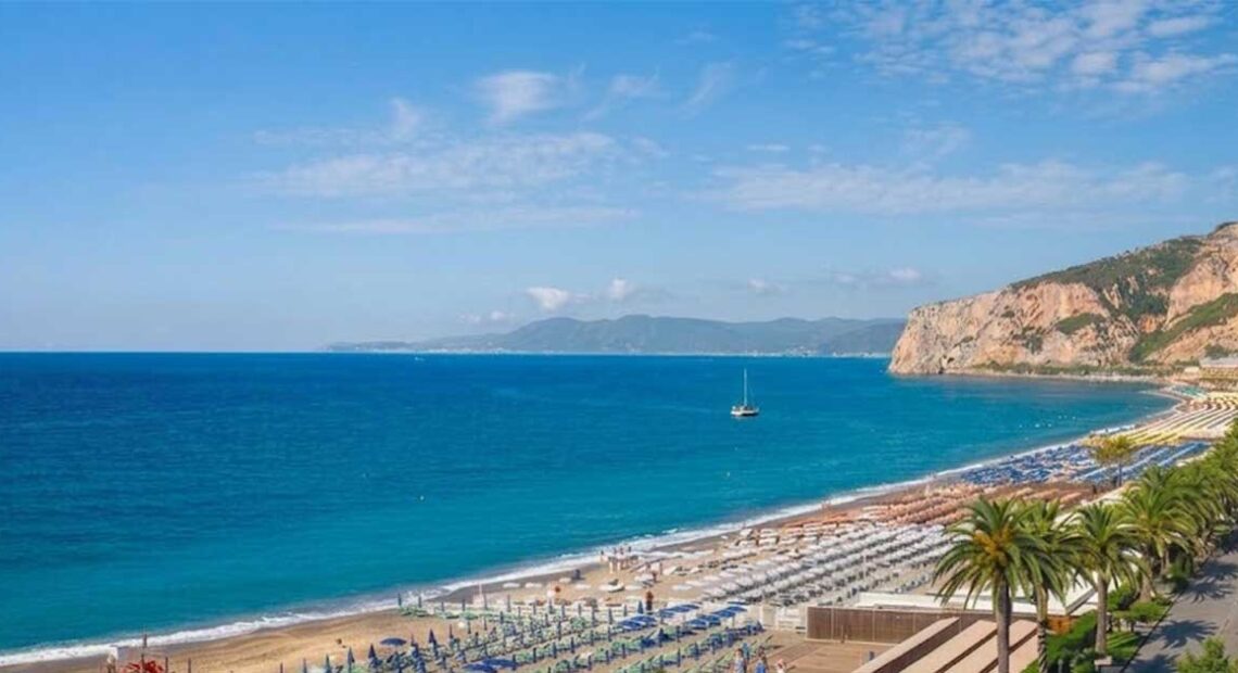 Panoramica dall'alto del litorale di Finale Ligure con gli stabilimenti balneari attrezzati, il lungomare con le palme e la falesia sullo sfondo.