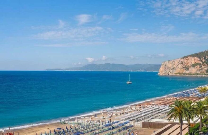 Panoramica dall'alto del litorale di Finale Ligure con gli stabilimenti balneari attrezzati, il lungomare con le palme e la falesia sullo sfondo.