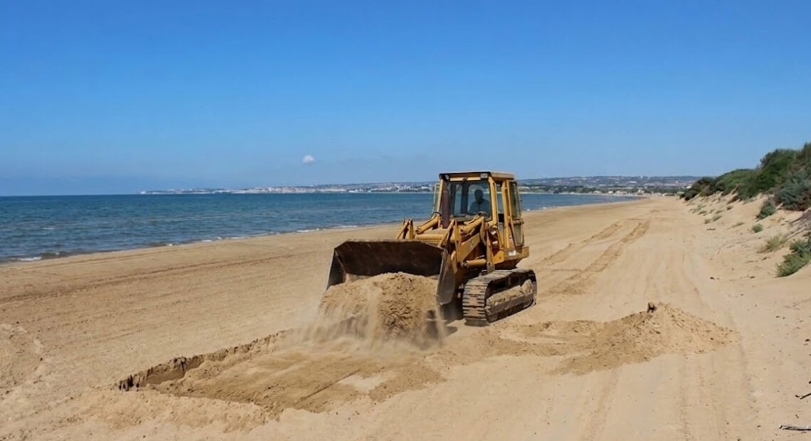 Un trattore cingolato in azione mentre solleva la sabbia su una spiaggia costiera della Sicilia, a ridosso delle dune e della vegetazione psammofila.