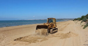 Un trattore cingolato in azione mentre solleva la sabbia su una spiaggia costiera della Sicilia, a ridosso delle dune e della vegetazione psammofila.