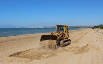 Un trattore cingolato in azione mentre solleva la sabbia su una spiaggia costiera della Sicilia, a ridosso delle dune e della vegetazione psammofila.