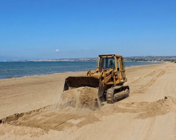 Un trattore cingolato in azione mentre solleva la sabbia su una spiaggia costiera della Sicilia, a ridosso delle dune e della vegetazione psammofila.