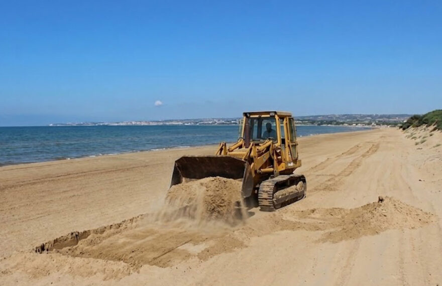 Un trattore cingolato in azione mentre solleva la sabbia su una spiaggia costiera della Sicilia, a ridosso delle dune e della vegetazione psammofila.