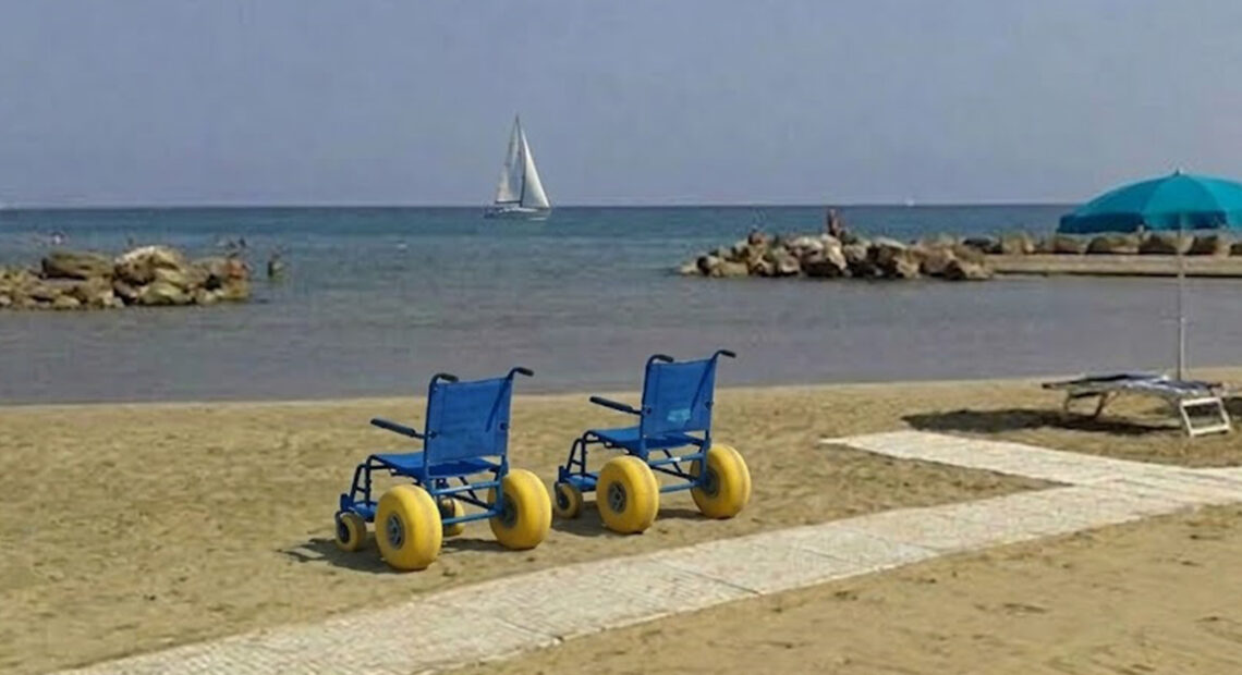 Due sedie JOB azzurre per la balneazione dei disabili posizionate sulla sabbia accanto a una passerella bianca, su una spiaggia attrezzata con ombrelloni e una barca a vela sullo sfondo.