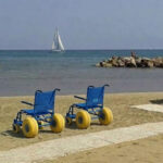 Due sedie JOB azzurre per la balneazione dei disabili posizionate sulla sabbia accanto a una passerella bianca, su una spiaggia attrezzata con ombrelloni e una barca a vela sullo sfondo.